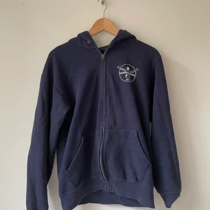 Blå zip-up hoodie - En lite vintage blå zip-up hoodie med vit tryck fram på och bak på. Hoodiens skicka är 7/10 då den är välanvänd med lite ytlig skada. Storleken är S och passformen är normal.