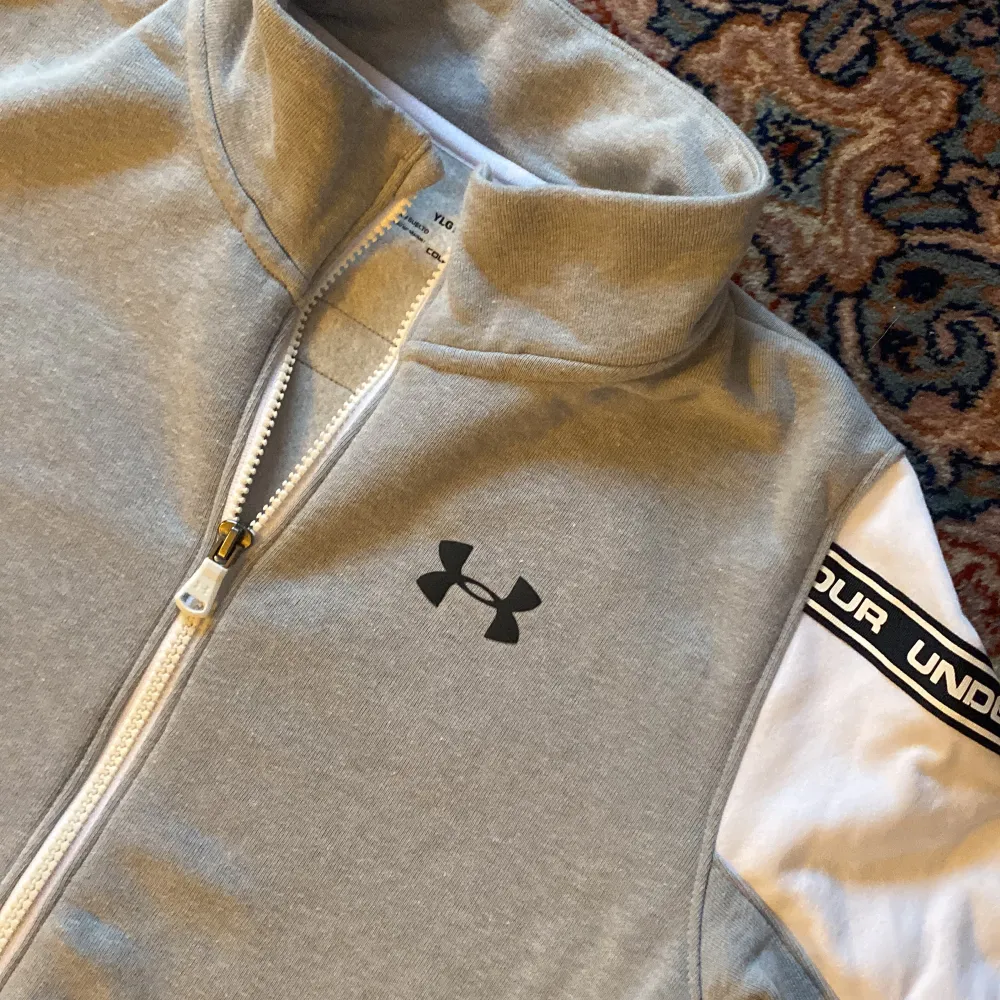 Långärmad Under Armour grå tröja med vita detaljer & dragkedja. Använd en gång. Skön insida i form av tunn fleece. Storlek anges ej, men    för barn ca 150 cm. Tröjor & Koftor.