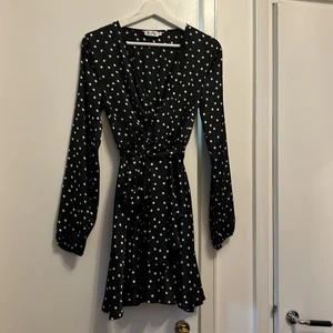 Omlottsydd satinklänning med volang - Svart satinklänning med vita prickar. V-ringad. Omlottsydd, knytband i midjan, resår i midjan. Volang i kjolen, liten smal mudd i ärmsluten. 