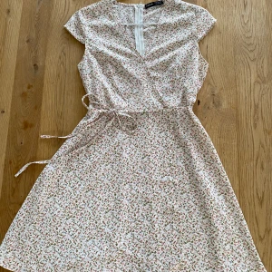 Klänning - Vit klänning med små rosa blommor från SHEIN. Storlek 34/XS. Snöret runt midjan går att stjustera, och även ta bort om man vill. Har aldrig kommit till användning, så är i nyskick!