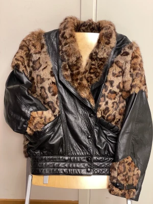Svart skinnjacka med leopardmönstrad kaninpäls - Grym svart skinnjacka med pälsdetaljer (leopardmönstrad kaninpäls). Ett unikt 80-talsplagg från spanska ”El Corte Inglés”. Fint svart quiltat foder. Stängs med tryckknappar. Märkt storlek 42. Jackan sitter på en provdocka med måtten 42 (längd ca 170cm). 