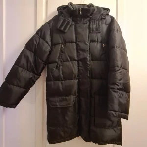 Svart lång vinterjacka  - Lång svart vinterjacka i nyskick, vattenavstötande och bra för en kall vinter. Två stora fickor med kardborreband. Dragkedja fram och extra skydd med knappar för att du ska hålla dig riktigt varm. Har luva. Endast använd ett par gånger.