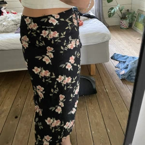 Kjol - Blommig kjol med hög slits, man kan knyta ett snöra som går runt midjan. Dragkedja vid slitsen. Slutar längre ner vid smalbenet. 