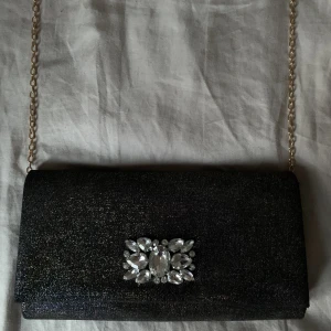 Glittrig axelväska  - Fin axelväska i svart glitter/skimmer. Strasstenar på framsidan och lång tunn guldkedja. Osäker på märke för fick den för några år sedan, men tror det är Glitter.  Aldrig använd så i nyskick🖤