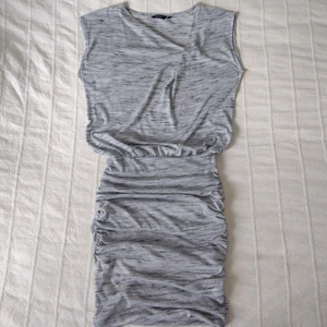 Gråmelerad, mjuk, stretchig klänning XS - Mjuk och stretchig klänning i en väldigt snygg och smickrande modell. Vit/svart/gråmelerad. En lösare överdel med fin "halsringning" och en tightare underdel med dubbla lager tyg och "krås" i sidorna. Sparsamt använd. 85% polyester och 15% viskos. Storlek XS.