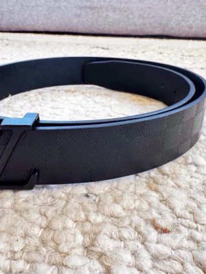 Svart Iv rutigt bälte - Snyggt svart bälte med diskret rutigt mönster och en stilren logga på metallspännet. Bältet är gjort i skinn och har en modern, minimalistisk look som passar till många outfits.