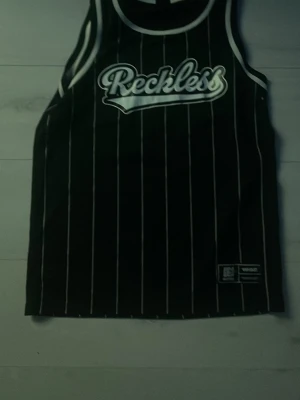 Svart randig linnetopp Reckless - Säljer en svart linnetopp från Reckless med vita vertikala ränder och vit logga framtill. Klassisk sportig look med ribbade kanter runt hals och ärmhål. Perfekt för dig som gillar streetwear och vill sticka ut.