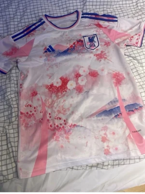 Japan landslagströja rosa/blå adidas - Säljer en Japan landslagströja från adidas i vitt med rosa och blå detaljer samt körsbärsblommor och bergsmotiv. Tröjan har blå ränder på axlarna och JFA-logga på bröstet. Perfekt för dig som gillar unika matchtröjor med snygg design.