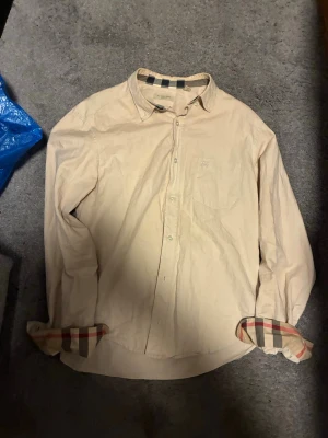 Burberry Skjorta - Säljer en beige skjorta från Burberry med klassiskt rutigt mönster på insidan av manschetterna och kragen. Skjortan har lång ärm, bröstficka med diskret broderad logga och knappar framtill. Perfekt för dig som gillar stilrena plagg med exklusiva detaljer. Vid byten eller fler frågor hör av er!
