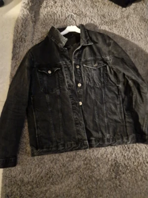 Svart jeansjacka med klassisk look - Säljer en svart jeansjacka med raka sömmar och två bröstfickor med knapp. Jackan har en klassisk krage och silverfärgade knappar framtill. Perfekt för dig som gillar en tidlös och avslappnad stil.