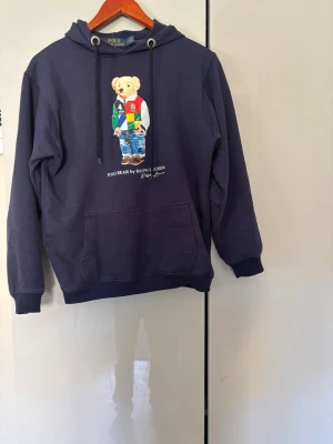 Marinblå hoodie Polo Ralph Lauren - Snygg marinblå hoodie från Polo Ralph Lauren med den ikoniska Polo Bear-trycket framtill. Hoodein är använd fåtal gånger. Hör gärna av dig om du har en fråga. Pris kan diskuteras.