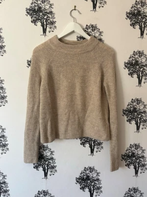 Beige stickad tröja med ribbad struktur - En beige stickad tröja med ribbad struktur och rund hals. Tröjan har långa ärmar och är tillverkad i ett mjukt material som ger en avslappnad och stilren look. Perfekt för lager-på-lager och passar till jeans eller kjol.