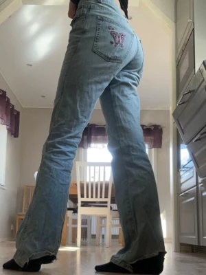 Blå bootcut jeans med fjäril - Säljer ett par ljusblå lågmidjade bootcut jeans med broderad rosa fjäril på bakfickan.💕Defekter syns på bilderna💕Midjemått: cirka 38 cm rakt över💕Passar mig som är 169 bra i längden 💕