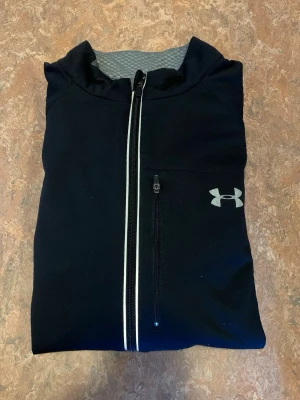 Svart träningsjacka från Under Armour - Snygg svart träningsjacka från Under Armour med hel dragkedja framtill och reflekterande detaljer. Jackan har en bröstficka med dragkedja och diskret logga på vänster sida. Insidan är grå och materialet känns lätt och funktionellt, perfekt för löpning eller gym.