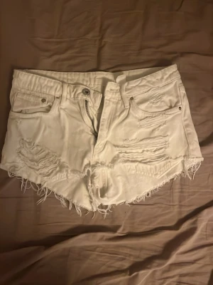 Vita fransiga jeansshorts - Säljer ett par vita jeansshorts med fransig kant och slitna detaljer framtill. Shortsen har fem fickor, knapp och dragkedja. Perfekta för sommaren och ger en avslappnad vibe. Low waist, och passar väldigt bra i midjan. Storlek 34.