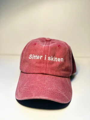 Keps ”sitter i skiten” röd  - Den där känslan när allt går åt helvete, men du ändå ser rätt bra ut. “Sitter i skiten” är kepsen för dig som har accepterat kaoset och gör det med stil. Lite självironi, lite swag – 100% du.  Tillverkad av 100% ekologisk bomull för maximal komfort, oavsett om du sitter i skiten på jobbet, i plugget eller i livet i allmänhet.