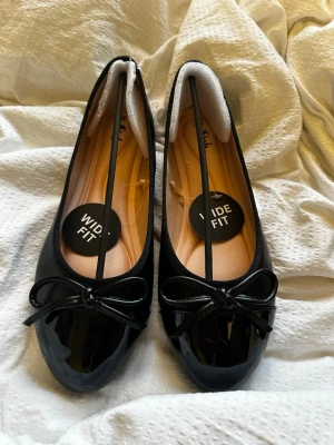 Svarta lackade ballerinaskor med rosett - Stilrena svarta ballerinaskor från Sole med glansig lackyta och söt rosettdetalj framtill. Modellen har bred passform (wide fit) och en mjuk, beige innersula för extra komfort. Perfekta för dig som vill ha både stil och bekvämlighet. W10 passar mig med storlek 41