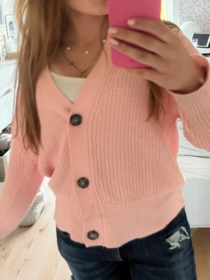 Ljusrosa stickad kofta med knappar - Säljer en ljusrosa stickad kofta med stora mörka knappar framtill. Koftan har v-ringning, ribbade muddar och en avslappnad passform. Perfekt att slänga över en topp för en söt och enkel look.