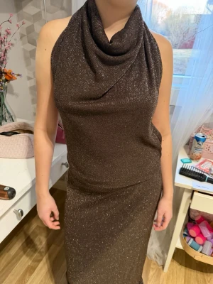 Brun glittrig balklänning - Säljer en brun maxiklänning med glittrigt tyg och halterneck. Klänningen har en draperad urringning och är figurnära med lång kjol. Perfekt för dig som vill sticka ut med en elegant look. Materialet är stretchigt och skimrar snyggt i ljuset.
