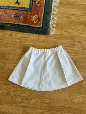 Vit kortkjol med knappar bak - Snygg vit kortkjol med utsvängd form och två dekorativa knappar bak i midjan. Kjolen har en enkel och stilren design som passar till många olika looks. Perfekt för dig som gillar minimalistisk stil.