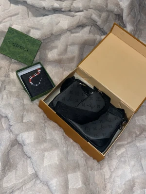 Louis vuitton trio väska + Gucci plånbok  - Snygg svart handväska från Louis Vuitton med klassiskt monogrammönster i mörkgrått. Väskan har bred axelrem, silverfärgade detaljer och en mindre avtagbar väska. Perfekt för dig som gillar exklusiva accessoarer och vill ha plats för det viktigaste. 