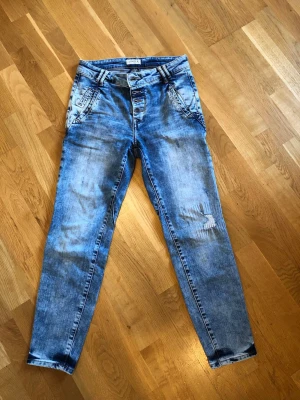 Blå jeans från Lindex, modell Maia  - Blå jeans från Lindex, modell MAIA. Jeansen har en tvättad look med slitningar på ena benet, knappgylf och fickor fram.