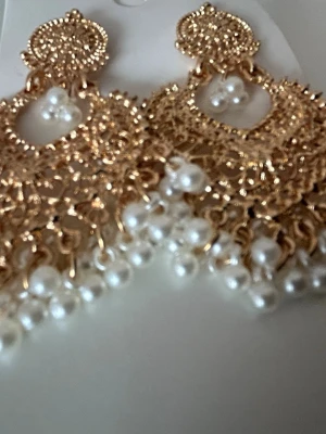 Guldpläterade örhängen med pärlor - Snygga och stora guldpläterade örhängen med intrikat filigranmönster och vita pärlor som hänger längs kanten. Örhängena har en elegant och lyxig design med flera små pärlor och detaljer som verkligen sticker ut.