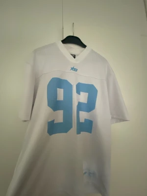 Vit t-shirt med blått tryck från FSBN - Säljer en vit t-shirt från FSBN med stort blått nummer 92 på framsidan. T-shirten har v-ringad hals och korta ärmar. Perfekt för dig som gillar sportig stil och vill sticka ut med en clean look.