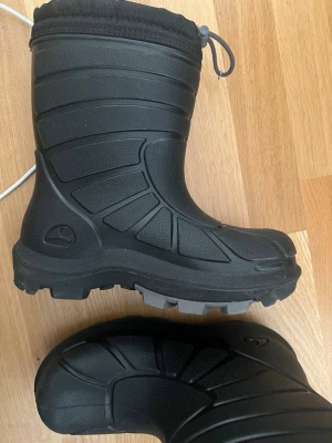 Svarta vinterstövlar med snölås - Säljer ett par svarta vinterstövlar med grov sula och snölås upptill. Insidan är fodrad med vitt, mjukt material för extra värme. Stövlarna har en sportig design och är perfekta för kalla dagar. Tillverkade i slitstark syntet och textil.