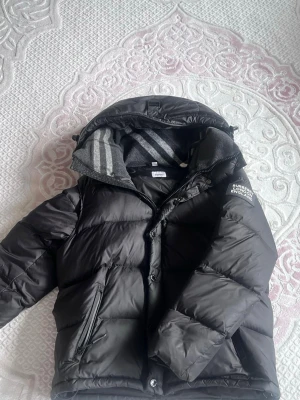 Svart dunjacka från Burberry - Snygg svart dunjacka från Burberry med huva och dragkedja. Jackan har en diskret logga på bröstet, flera fickor och quiltad design. Perfekt för kalla vinterdagar och riktigt varm tack vare dunfyllningen. Klassisk och stilren look.