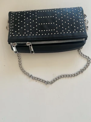 Svart axelväska med nitar och kedja - Snygg svart axelväska i konstläder med silverfärgade nitar på locket och en  silverkedja som axelrem. Väskan har en dragkedja framtill och coola detaljer som ger en cool vibe. Perfekt för dig som gillar axelväskor och piffa till din outfit! Väskan är aldrig använts. Den är köpt på Zalando och är ifrån märket even & odd 