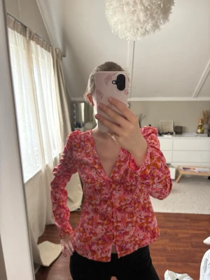 Blommig rosa blus med v-ringning - Säljer en färgglad blus med blommigt mönster i rosa, rött och orange toner. Blusen har v-ringning, figurnära passform och långa ärmar med lätt puff vid axlarna. Perfekt för dig som gillar färg och mönster!
