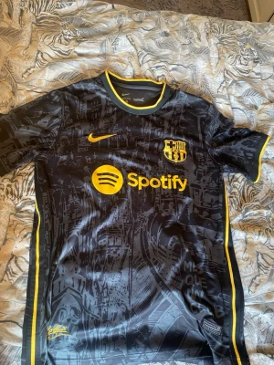 Svart FC Barcelona matchtröja Nike - Snygg svart FC Barcelona matchtröja från Nike med gula detaljer och tryck. Tröjan har klubbmärke, Spotify-logga och UNHCR-tryck samt ett unikt mönster inspirerat av Barcelona. Tillverkad i ett lätt och ventilerande funktionsmaterial, perfekt för fotboll eller träning.
