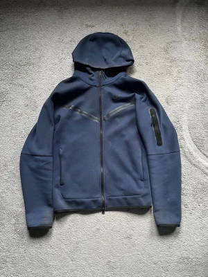 Nike tech fleece - Snygg mörkblå hoodie från Nike med dragkedja framtill och huva. Hoodien har två fickor fram och en extra ficka med dragkedja på ena ärmen. Tillverkad i ett tjockare, mjukt material som känns sportigt och modernt. 