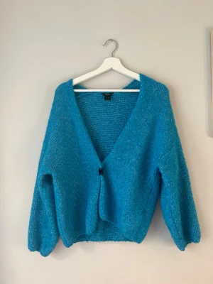 Blå fluffig kofta med v-ringning - Säljer en blå, fluffig kofta med djup v-ringning och ballongärmar. Koftan har en enkel knäppning framtill och är stickad i ett mjukt, glansigt material som ger en mysig känsla. Perfekt för att addera färg till din outfit.