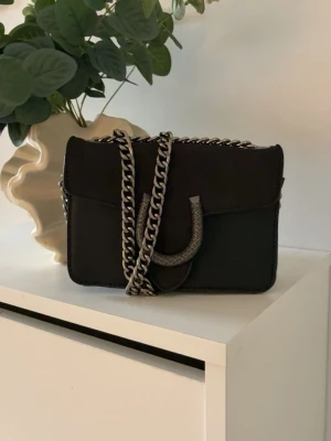 Svart handväska  - Snygg svart handväska från gunnes väskor med en hästsko som märke.silverfärgad kedjerem och dekorativt spänne framtill. Väskan har en boxig form och är tillverkad i en mix av mocka och läder. Perfekt accessoar för att lyfta din outfit.