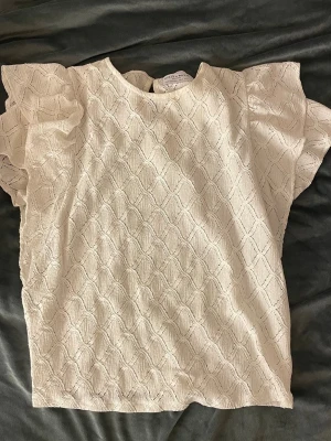 Vit blus från Holly & Whyte - Säljer en söt vit blus från Holly & Whyte med strukturmönster och volangärmar. Blusen har rund halsringning och knapp i nacken. Perfekt till jeans eller kjol för en fräsch look.