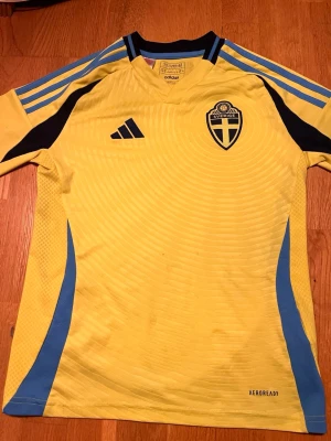 Svensk landslagströja Adidas gul/blå - Gul svensk landslagströja från Adidas med blå detaljer och svarta inslag. Tröjan har broderad landslagssköld, Adidas-logga och klassiska ränder på axlarna. Tillverkad i lätt funktionsmaterial (AEROREADY) som andas, perfekt för fotbollsträning eller match.