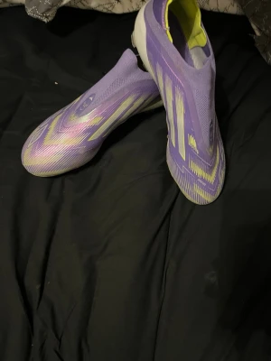 Lila Adidas X Speedflow fotbollsskor - Säljer ett par lila Adidas X Speedflow fotbollsskor med gula och vita detaljer. Skorna har en strömlinjeformad design utan snörning och ovandel i syntetmaterial för lätt vikt och bra bollkontroll. Perfekta för dig som vill sticka ut på planen.