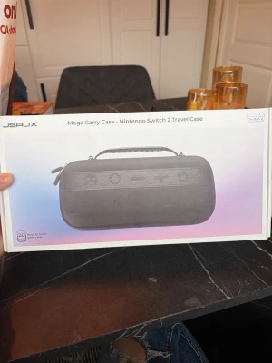 JSaux Mega Carry Case till Switch 2 - Snygg och praktisk svart väska från JSaux för Nintendo Switch 2. Väskan är tillverkad i slitstarkt material och har ett handtag för enkel transport. Perfekt för att förvara och skydda din konsol och tillbehör när du är på språng.