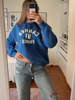 Blå sweatshirt - Blå sweatshirt med vit text 'CANNES 91 FRANCE' på bröstet. Tröjan har en avslappnad passform, rund halsringning och ribbade muddar vid ärmar och nederkant. Perfekt för en chill och sportig look.
