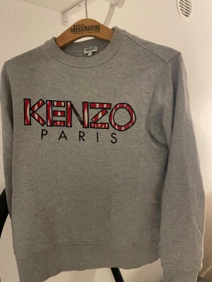 Grå Kenzo sweatshirt med tryck - Säljer en grå sweatshirt från Kenzo med stort, färgglatt Kenzo Paris-tryck i rött, vitt och svart på bröstet. Tröjan har rund halsringning, ribbade muddar och är tillverkad i mjuk bomullsblandning. Perfekt för en avslappnad och trendig look. passar till strl  (S-m)