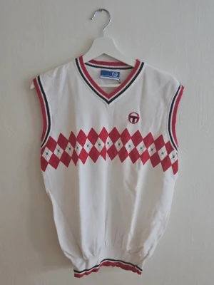 Vit och röd väst från Sergio Tacchini - Klassisk ärmlös väst från Sergio Tacchini i vitt med röd och marinblå argylemönster över bröstet. V-ringad med ribbade kanter i rött och blått. Perfekt för en sportig och retro look.