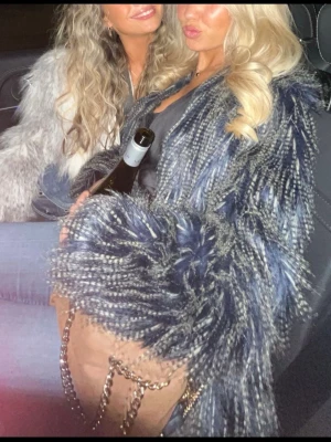 Blå-svart fluffig pälsjacka - Säljer en supercool blå-svart pälsjacka med lång, fluffig fuskpäls och öppen front. Jackan har en unik textur med inslag av vitt och ger en riktigt trendig look. Perfekt för dig som vill sticka ut och hålla dig varm. Passar till både vardags och fest. 