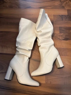 Vita slouchy stövlar med klack - Säljer ett par trendiga vita stövlar med hög klack och slouchy skaft. De har spetsig tå och bred blockklack för extra attityd. Under knäna. Tillverkade i ett läderliknande material som ger en clean och modern look. Perfekta för dig som vill sticka ut med din outfit.