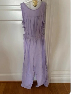 Ljuslila ärmlös jumpsuit - Säljer en ljuslila ärmlös jumpsuit med vida ben och resår i midjan. Jumpsuiten har dekorativa knytband på sidorna och är tillverkad i ett mjukt, luftigt tyg som känns skönt mot huden. Perfekt för dig som gillar en avslappnad och trendig stil.