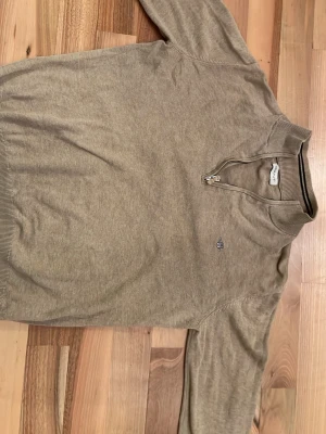 Beige halvzip-tröja från Gant - Klassisk beige tröja från Gant med halv dragkedja och liten broderad logga på bröstet. Tröjan har ribbade muddar vid ärmslut och nederkant samt hög krage. Perfekt för en avslappnad men ändå stilren look.