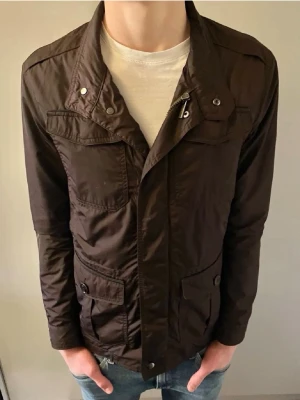 Field jacka från Massimo Dutti - Stilren  jacka från Massimo Dutti med flera fickor framtill och knappdetaljer. Jackan har lång ärm, normal passform och dragkedja samt tryckknappar framtill. Perfekt för dig som gillar en klassisk look med moderna detaljer.