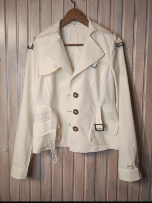 Vit trenchcoat från Tommy Hilfiger - Stilren vit trenchcoat från Tommy Hilfiger med stora knappar framtill, klassisk krage och dekorativa axelremmar. Jackan har två fickor med lock, bältesdetaljer och knappslå vid ärmsluten. Tillverkad i ett lätt bomullsmaterial, perfekt för vår och höst.