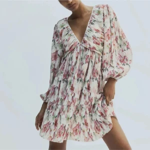 Blommig klänning med v-ringning - Superfin kortklänning med volanger och djup v-ringning. Klänningen är vit med rosa och gröna blommor, har långa puffärmar och är tillverkad i ett lätt, plisserat material. Helt i nyskick⭐️❤️